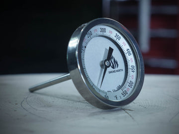 A Tel Tru offset smoker thermometer with custom 'Smoke North' logo shown on top of a wood splitting log